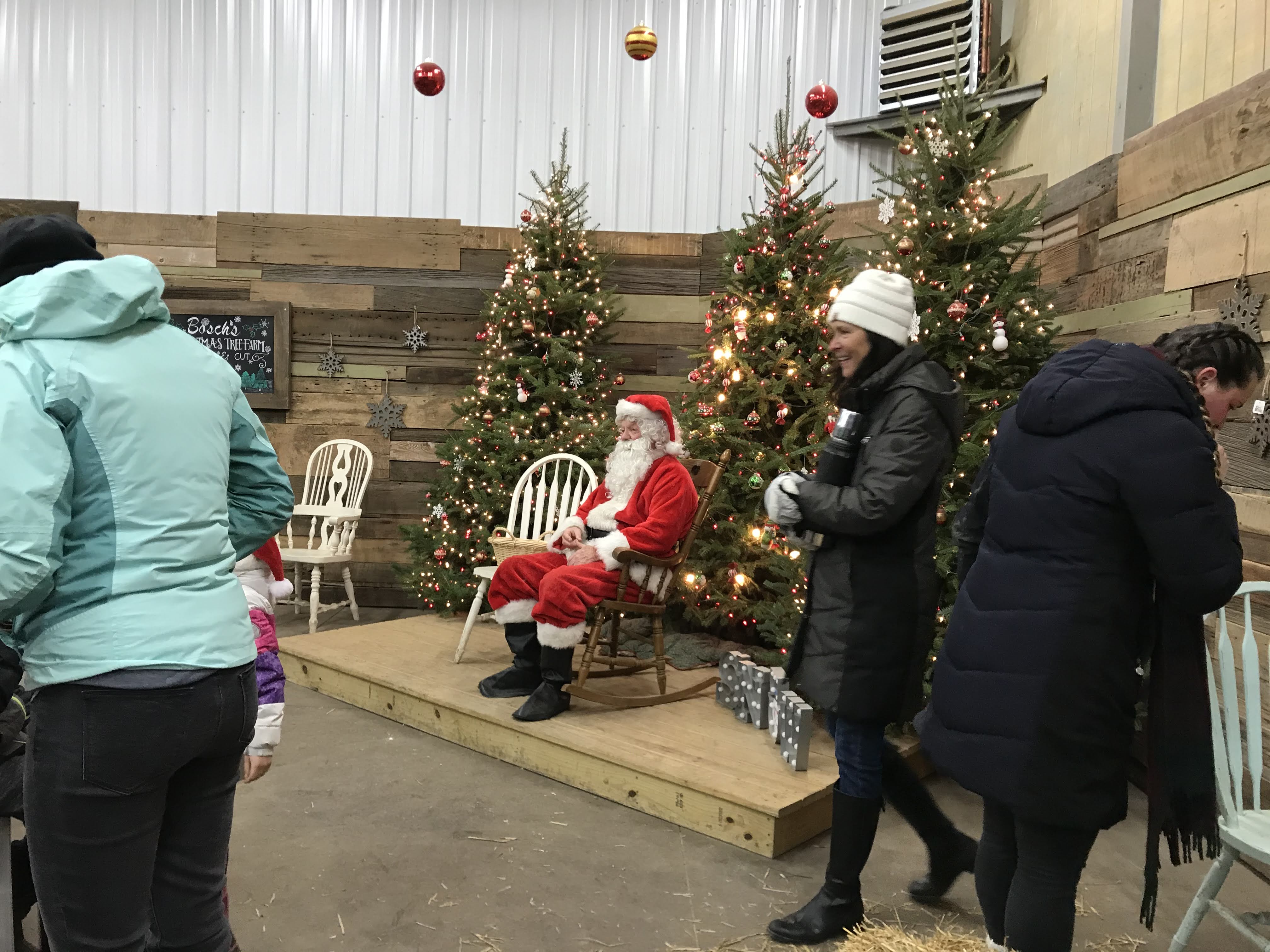 Santa sitting in a wooden rocking chair in front of lit up Christmas trees.